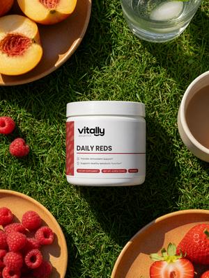A white jar of Vitally Essential Daily Reds dietary supplement placed on green grass, surrounded by fresh raspberries, sliced peaches, strawberries, and a glass of water in a styled outdoor flat-lay composition.
