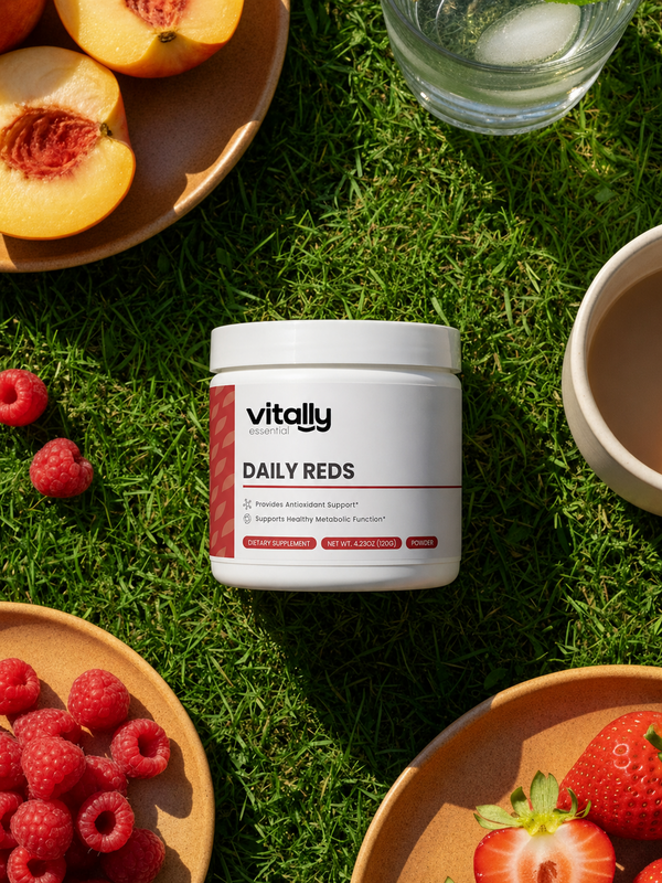 A white jar of Vitally Essential Daily Reds dietary supplement placed on green grass, surrounded by fresh raspberries, sliced peaches, strawberries, and a glass of water in a styled outdoor flat-lay composition.