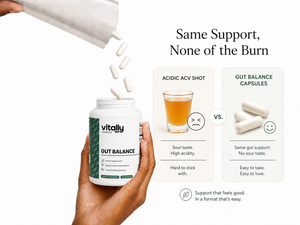 A hand holds a white bottle labeled 'Vitally Essential GUT BALANCE' while capsules pour out from an overturned white package. The right side includes marketing text comparing acid-based shots with gut balance capsules, with illustrated cups and capsules on a clean white background.