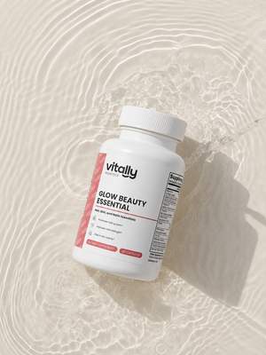 A white bottle of Vitally Essential Glow Beauty Essential dietary supplements, styled on a light beige surface with water ripple patterns and a splash behind it.