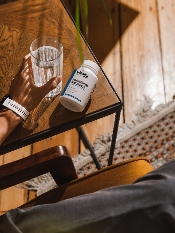 A white bottle of Vitally Essential Magnesium Oxide supplements with blue text, resting on a wooden table next to a glass of water, with a person's hand and arm visible in a warm, rustic indoor setting.